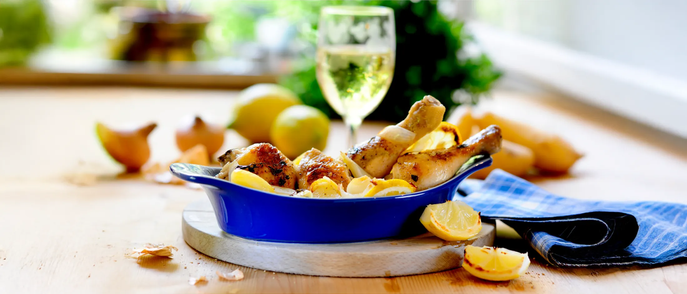 Roasted chicken drumsticks with lemon slices in a blue dish, served on a wooden board with a glass of white wine in the background.