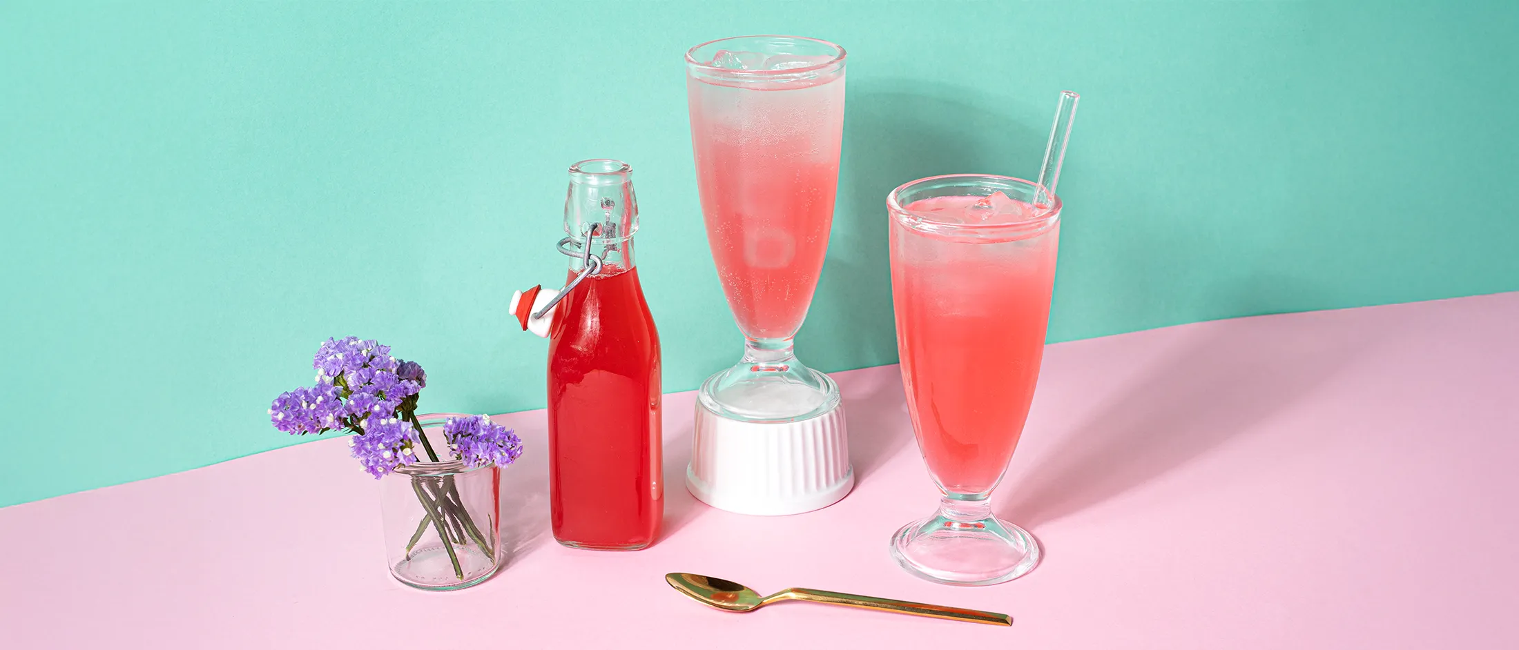 Three pink beverages in different containers, a small vase with purple flowers, and a gold spoon on a pink and teal background.