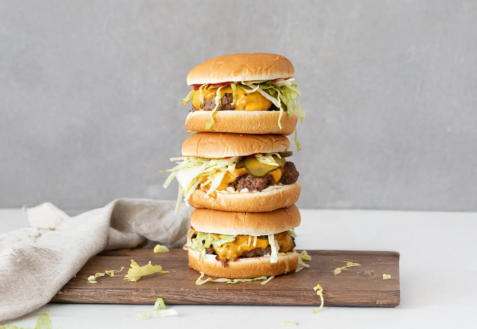 Three stacked cheeseburgers with lettuce, pickles, and melted cheddar cheese on a wooden board against a gray background.