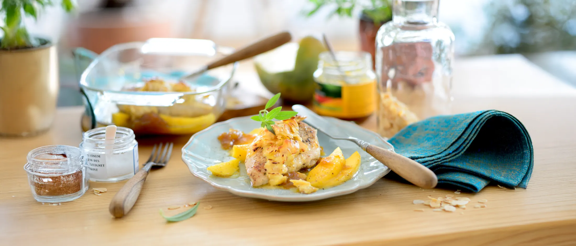 Plate of cooked fish topped with fruit and herbs, surrounded by utensils, a glass container, jar, and blue cloth on a wooden table.