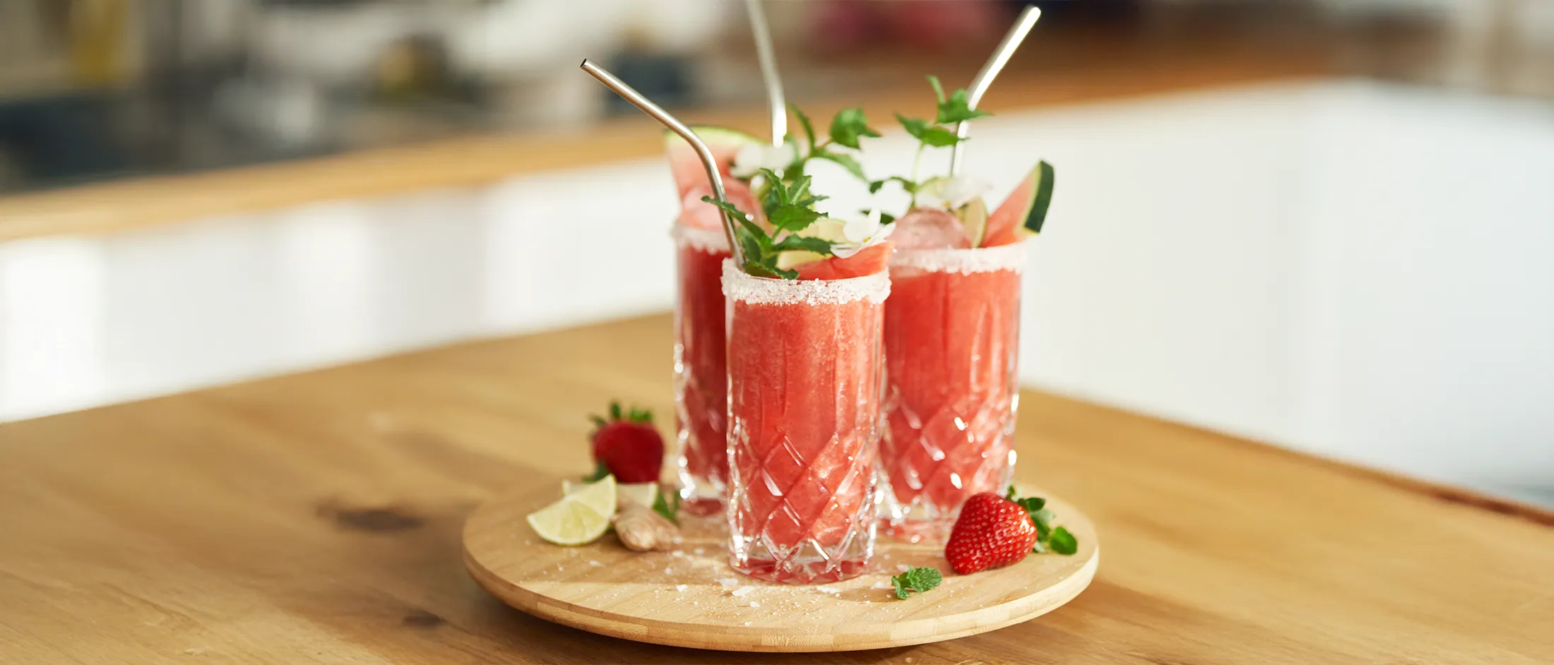 Three tall glasses of watermelon smoothie garnished with mint, lime, and salt rims, placed on a wooden tray with fresh fruits around.