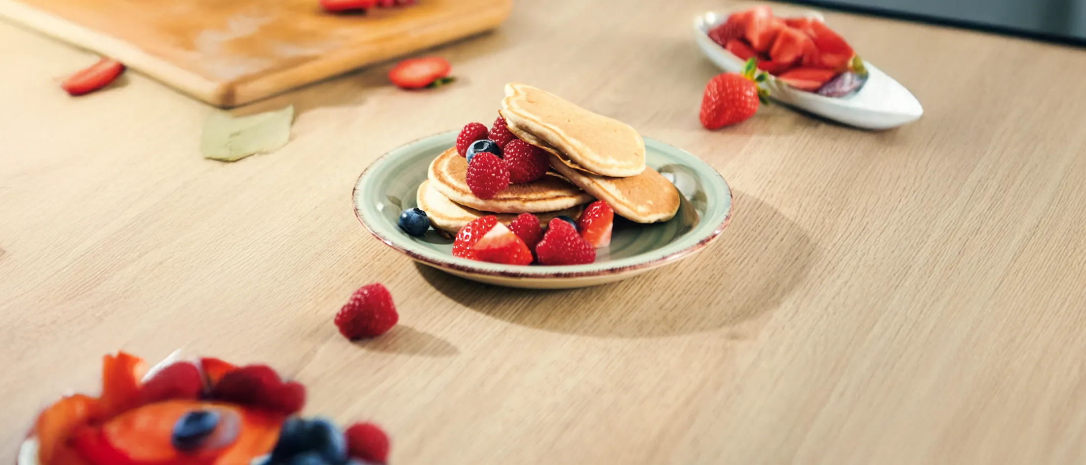 Stack of three golden pancakes on a plate topped with fresh raspberries, strawberries, and blueberries on a wooden table.