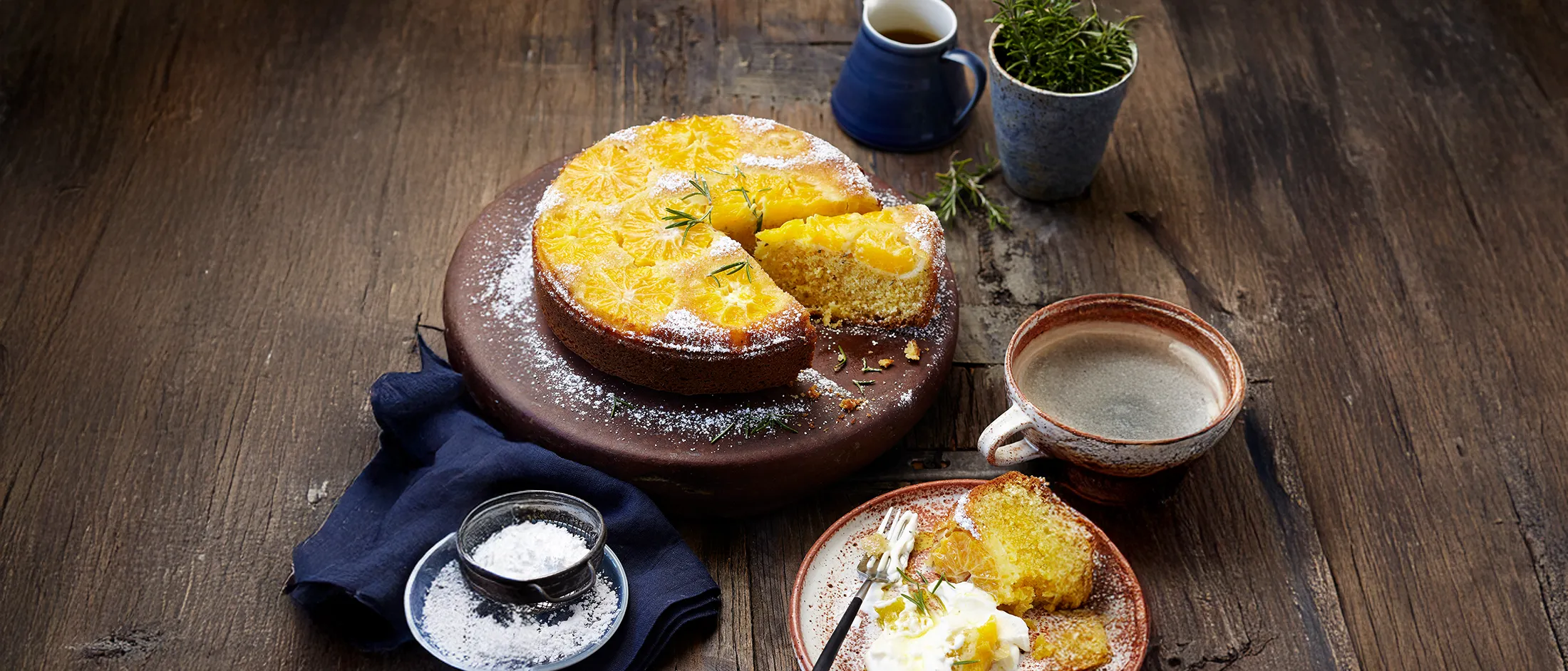 A sliced orange upside-down cake dusted with powdered sugar, served with cream on a plate, accompanied by coffee on a rustic wooden table.