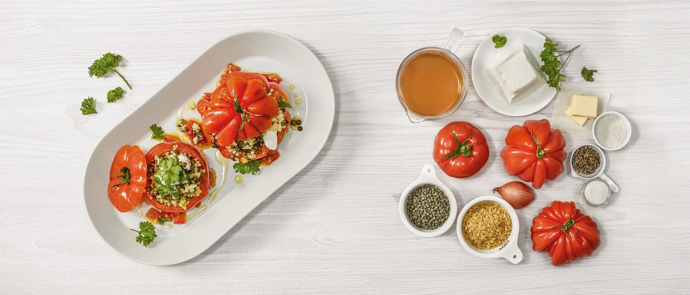 Plate with two stuffed tomatoes filled with grains and herbs next to ingredients including tomatoes, lentils, rice, broth, and cheese on white table.