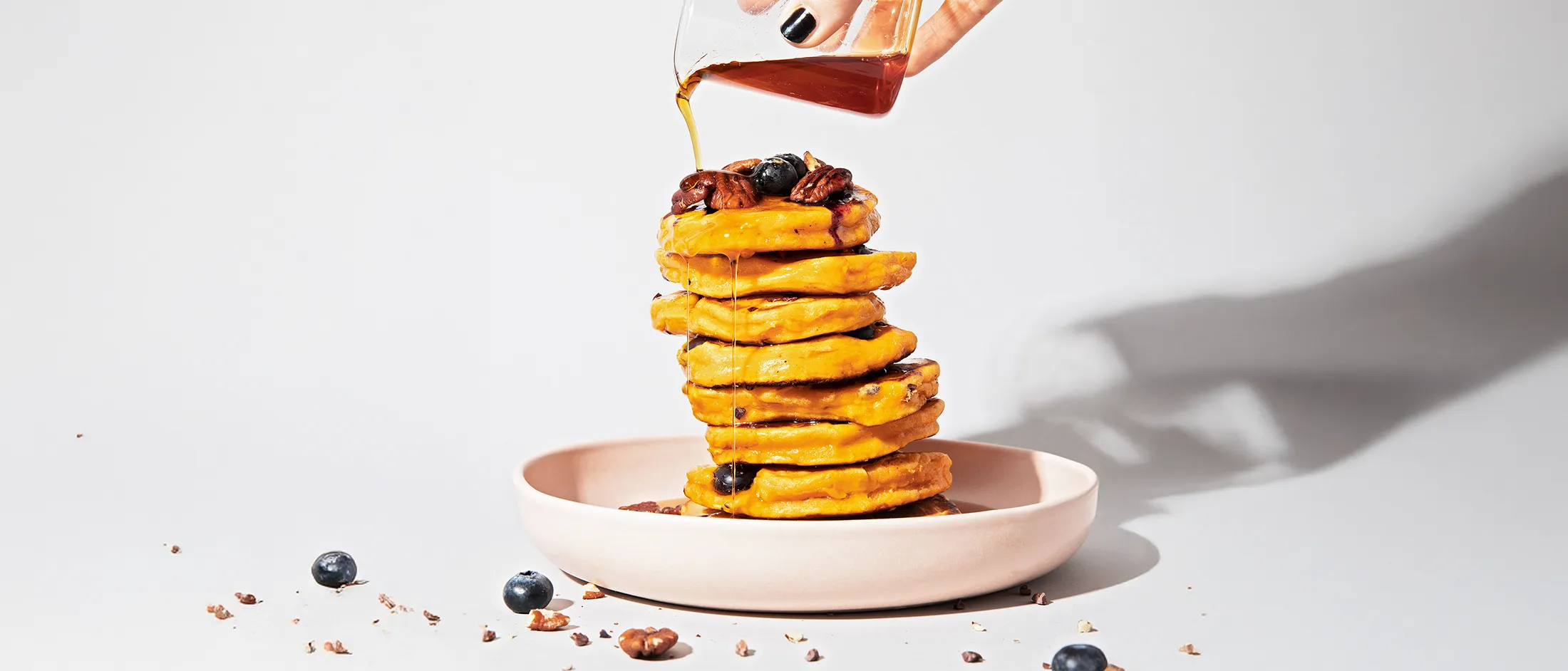 Stack of pumpkin pancakes topped with pecans and blueberries, with syrup being poured over them on a light pink plate.