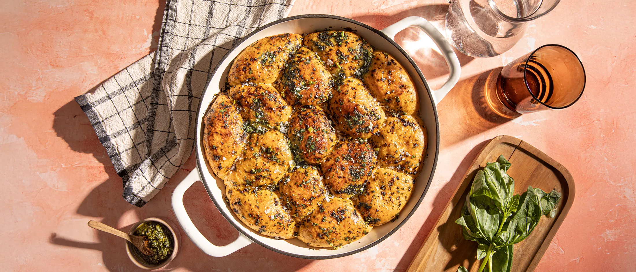 Golden brown herb-seasoned rolls in a round white baking dish, with coarse salt sprinkled on top, surrounded by herbs and glassware.