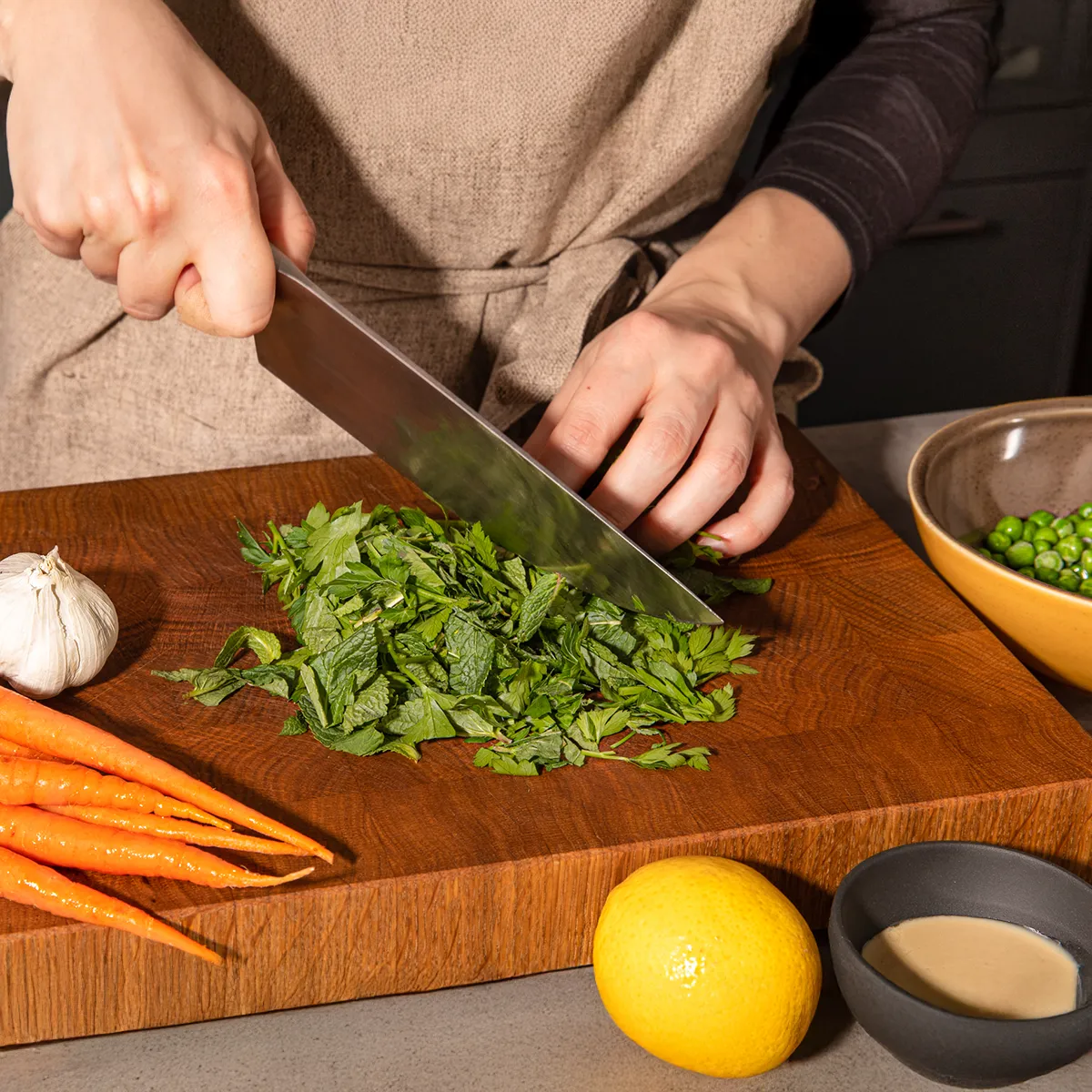 A person in an apron is finely chopping fresh herbs on a wooden cutting board, with carrots, garlic, and a lemon nearby.