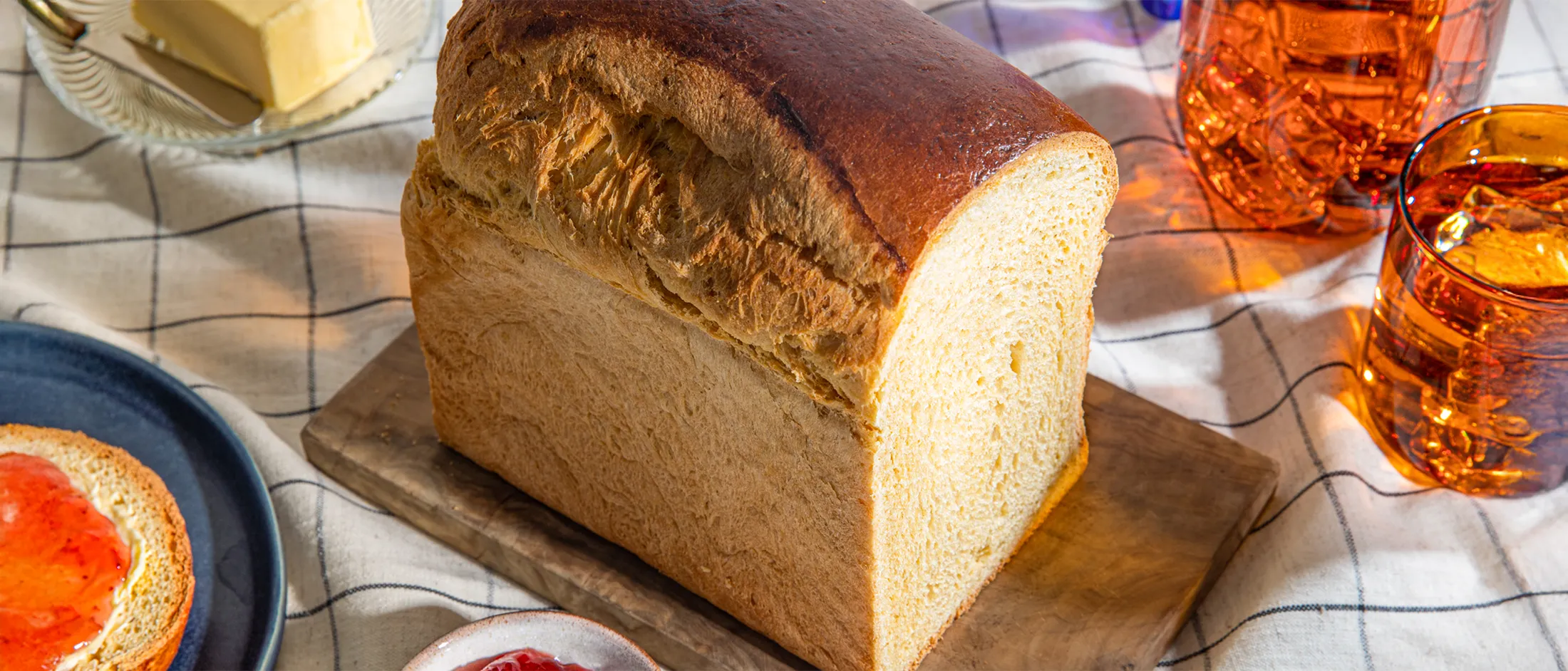 Freshly baked loaf of bread, butter, jam, and a glass of amber-colored liquid on a checkered tablecloth.