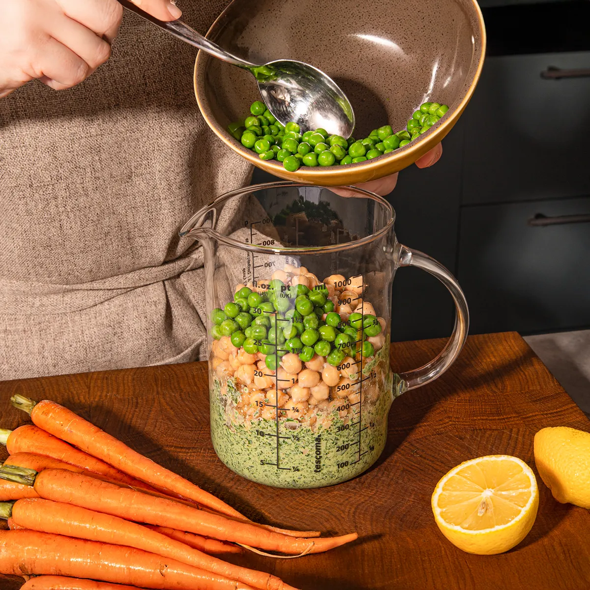 A person is adding green peas from a bowl into a large measuring jug containing chickpeas and a green mixture, with carrots and lemons on the wooden surface.
