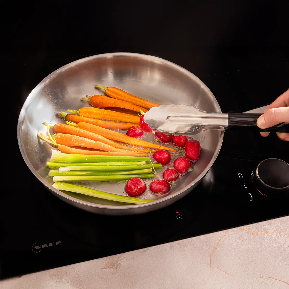 A hand uses tongs to turn vegetables in a stainless steel pan on a black induction cooktop.