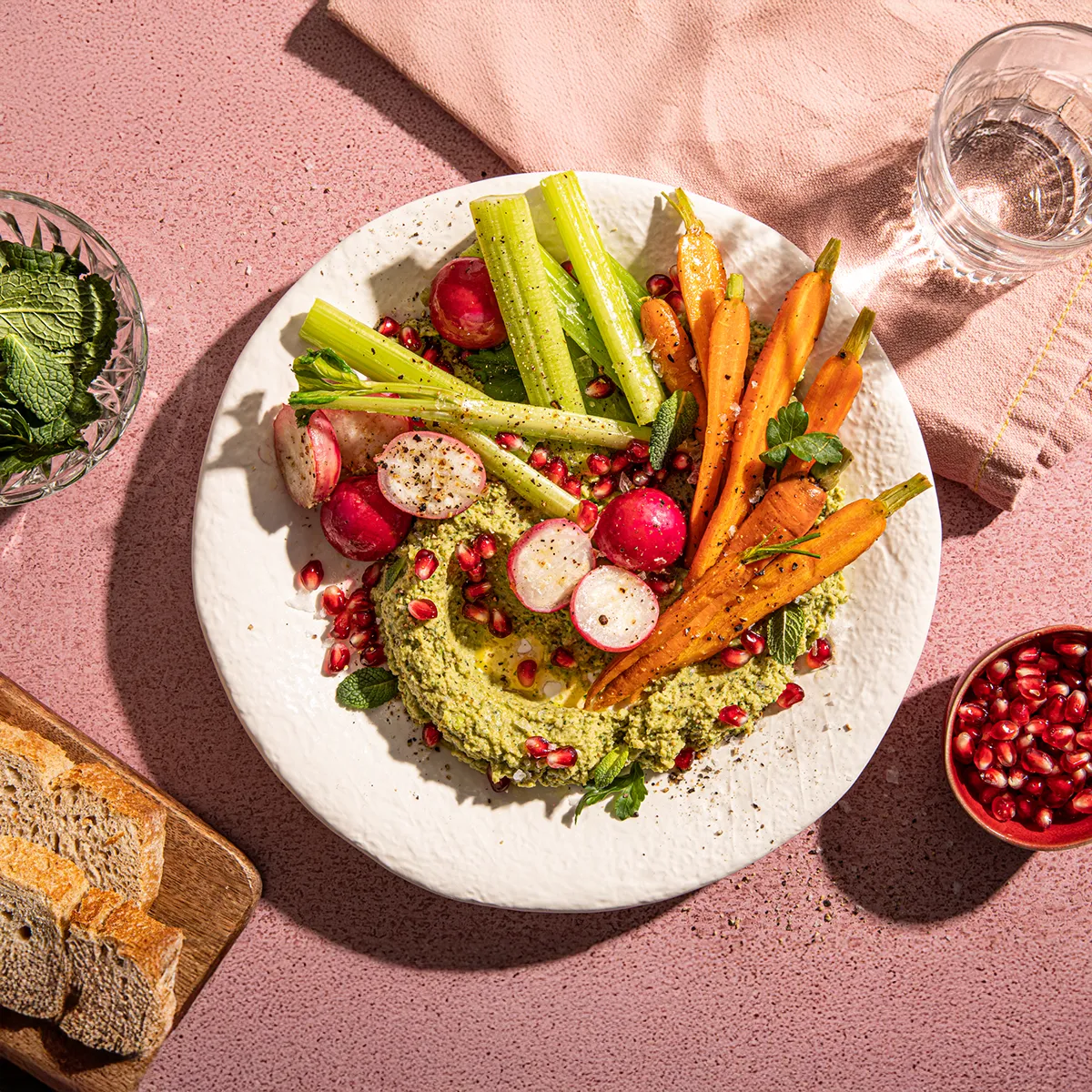 A vibrant platter of green hummus, topped with roasted carrots, radishes, celery sticks, and pomegranate seeds.
