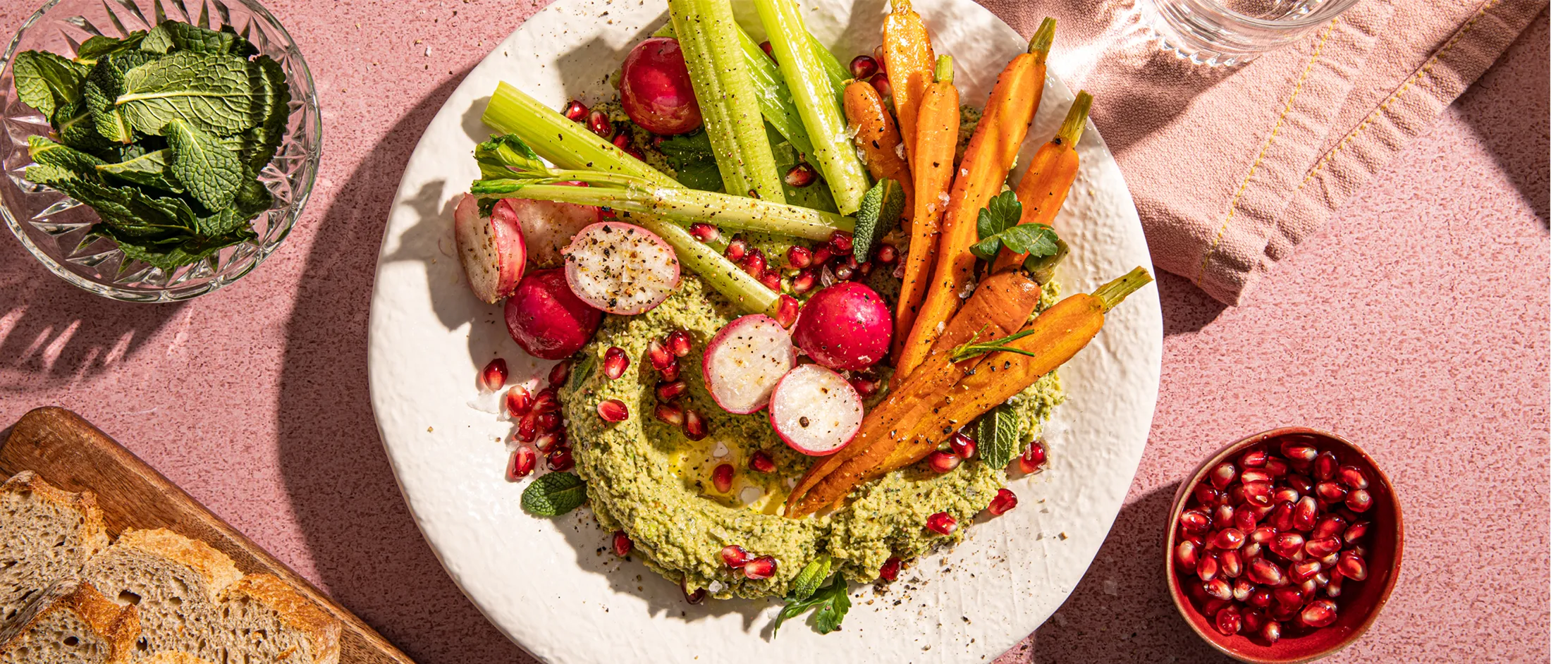 Plate of green hummus topped with radishes, baby carrots, celery sticks, and pomegranate seeds, with bread and mint garnish nearby.