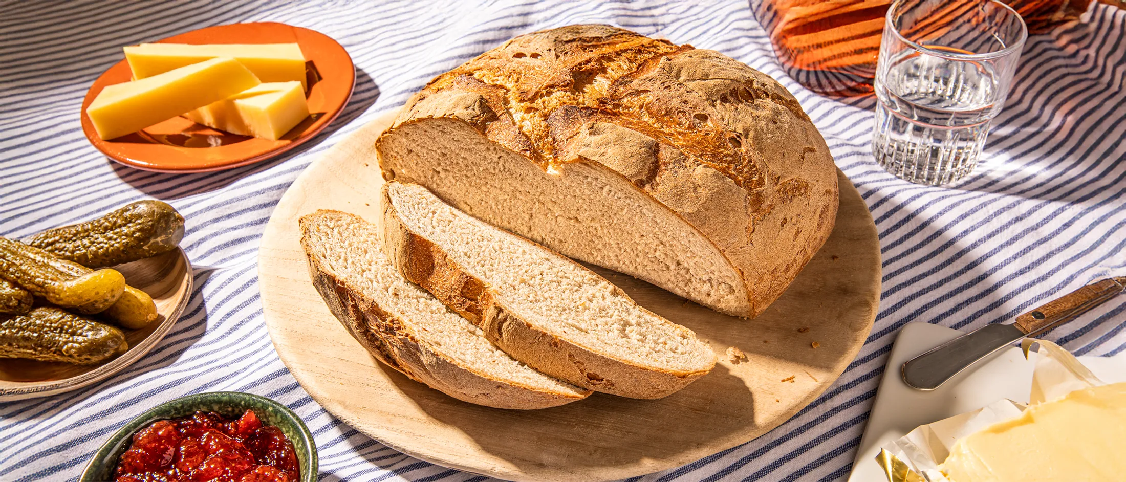 Round rustic loaf of bread sliced on a wooden board, surrounded by cheese, pickles, jam, butter, and a glass of water on a striped cloth.