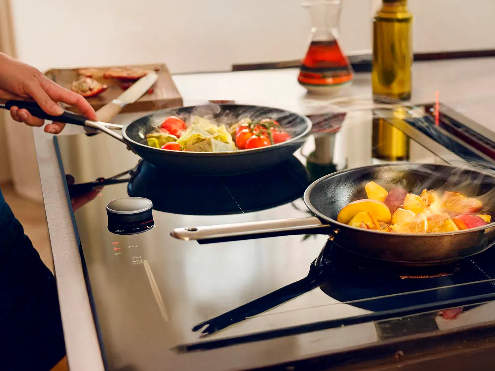 Due padelle su un moderno piano cottura con cibo fumante in preparazione, inclusi pomodori e frutta a fette, con prodotti freschi sul bancone nelle vicinanze.