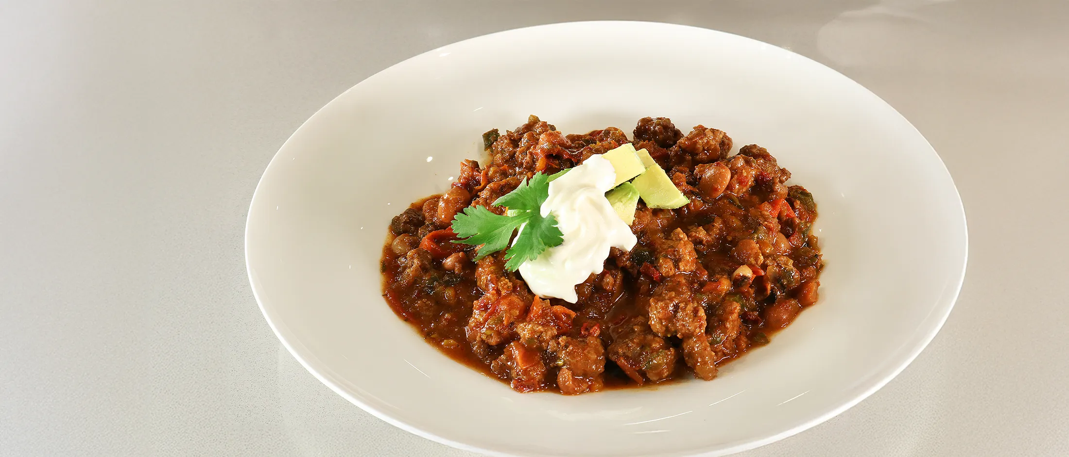 Bowl of chili topped with sour cream, avocado slices, and a cilantro leaf on a light surface.