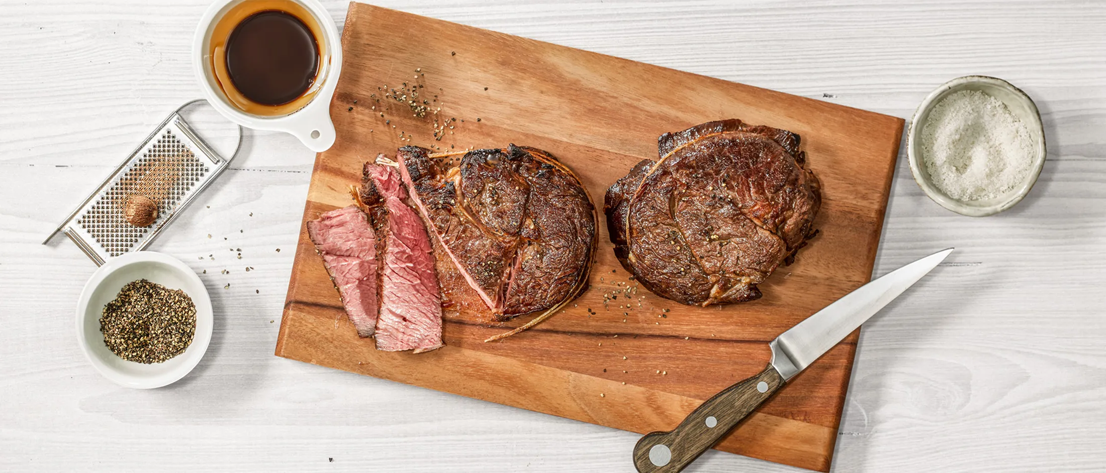 Two cooked ribeye steaks on a wooden board with slices cut, a knife, and a cup of black coffee.