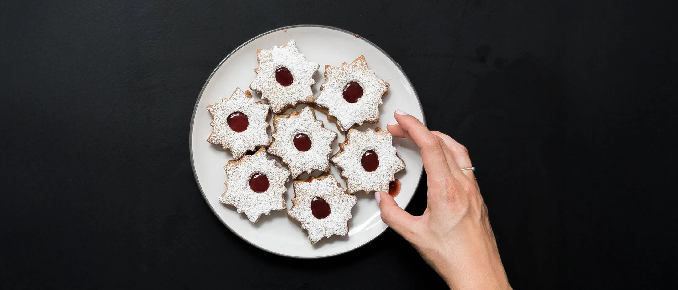 Hand reaching for a jam-filled linzer cookie dusted with powdered sugar on a white plate with seven cookies.