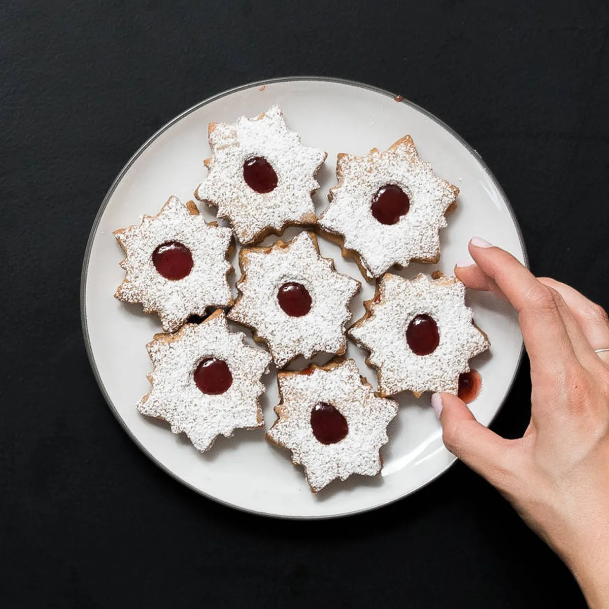 Hand reaching for a jam-filled linzer cookie dusted with powdered sugar on a white plate with seven cookies.