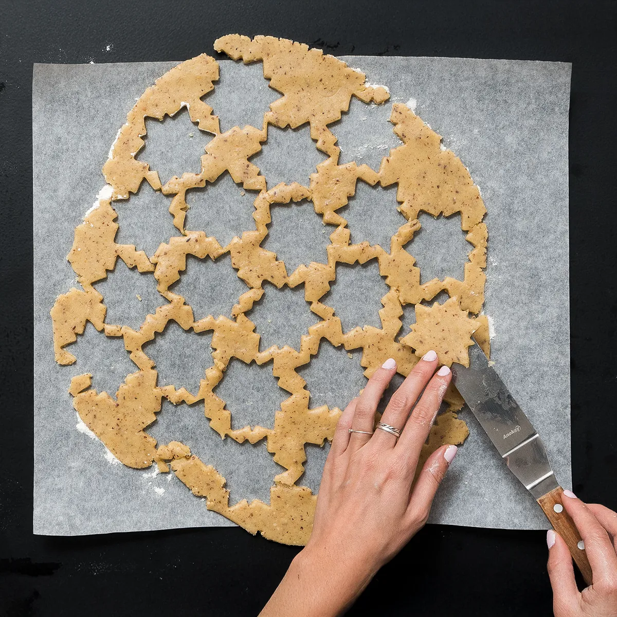 Hand removing star-shaped cookie dough cutouts from parchment paper with a spatula on a dark surface.