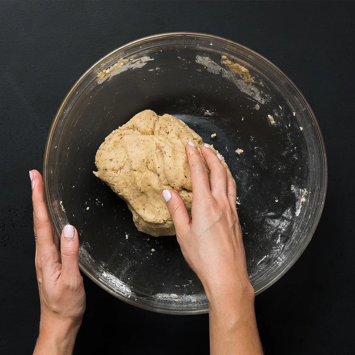 Hands kneading dough in a clear glass bowl on a black surface, with some flour scattered inside the bowl.