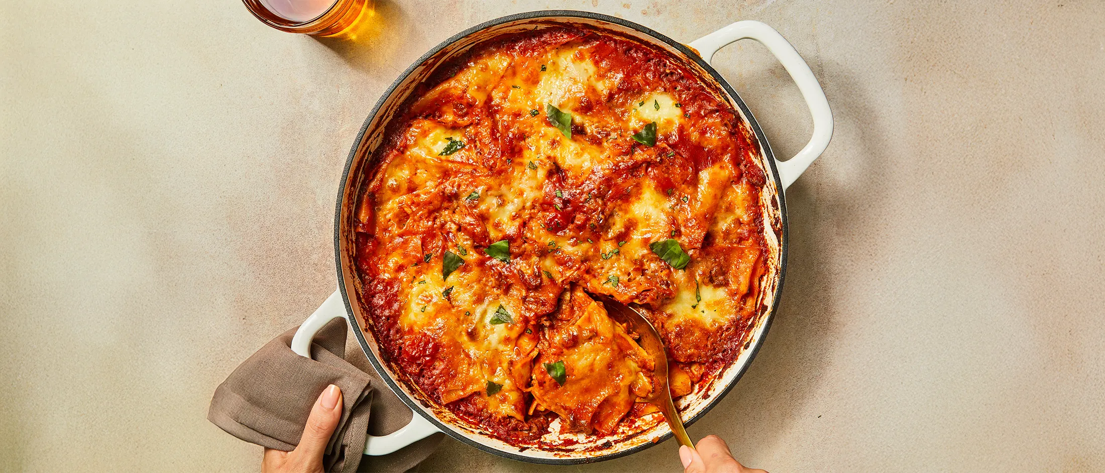 Pan of baked lasagna topped with melted cheese and fresh basil, with a person serving a portion using a spoon.