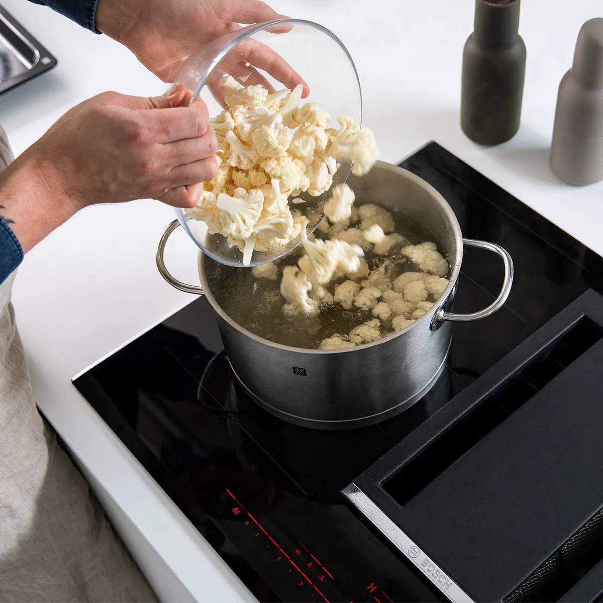 Person adding cauliflower florets from a glass bowl into a pot of boiling water on a stovetop.