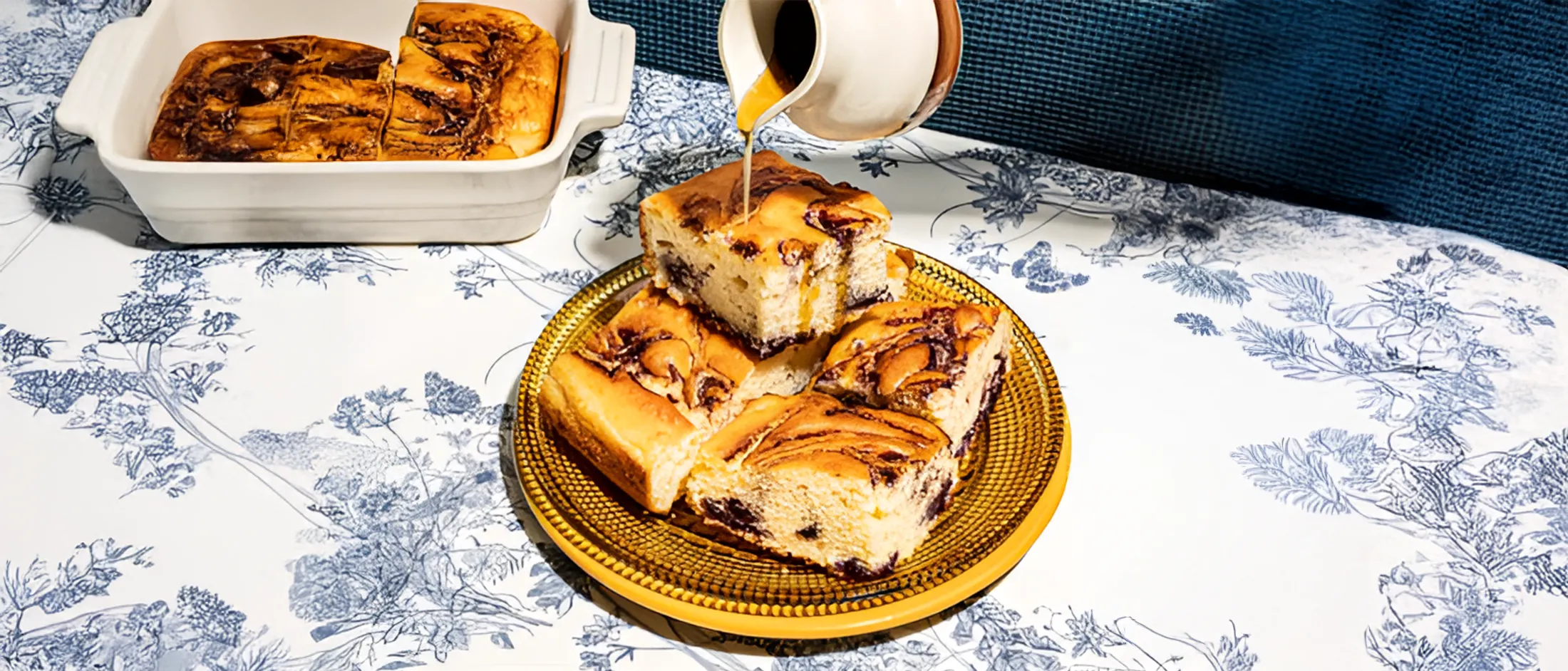 A golden plate holds several squares of swirled coffee cake, with a pitcher pouring syrup over them, and a baking dish in the background.