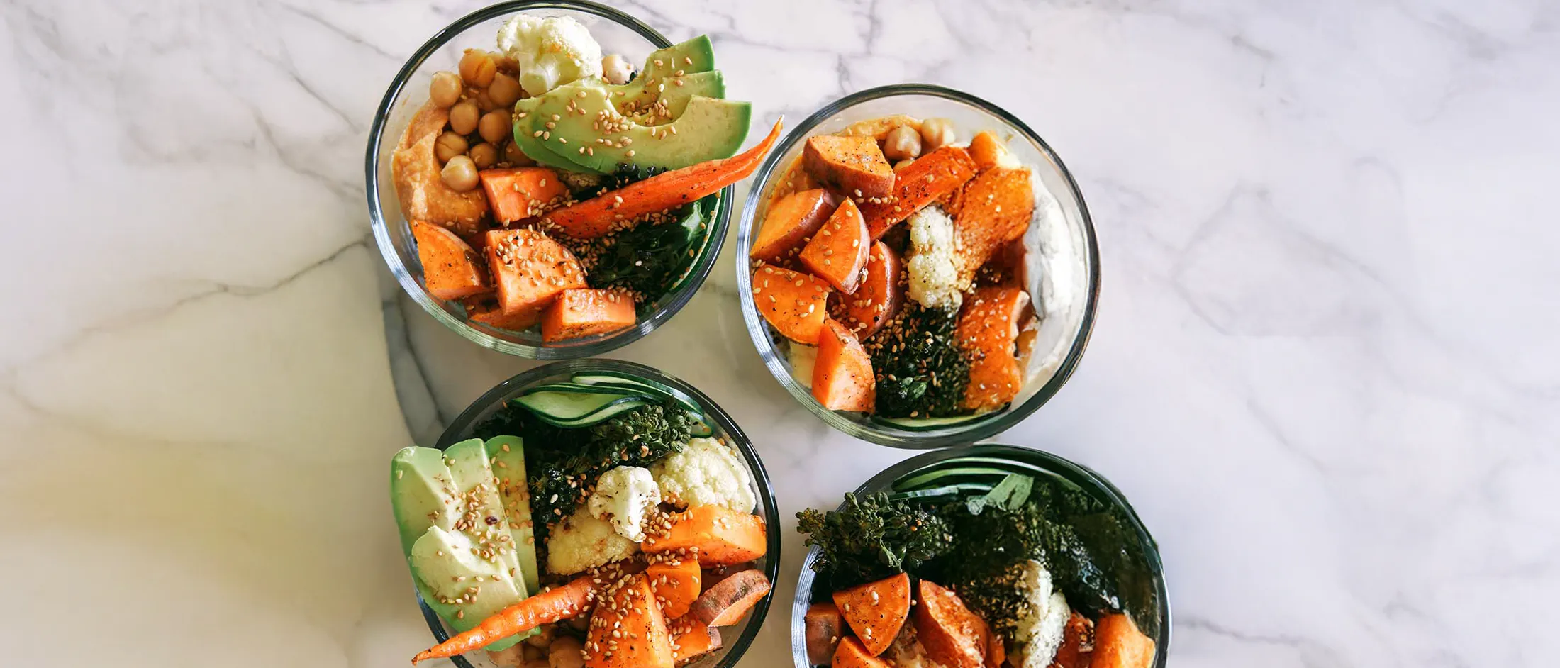 Top-down view of four glass meal‑prep bowls arranged on a white marble countertop, each filled with roasted sweet potato chunks and whole baby carrots, chickpeas, sliced avocado, cauliflower florets and wilted greens, all sprinkled with sesame seeds; one bowl has a pale dollop of creamy spread and a stack of teal plastic lids sits at the edge.