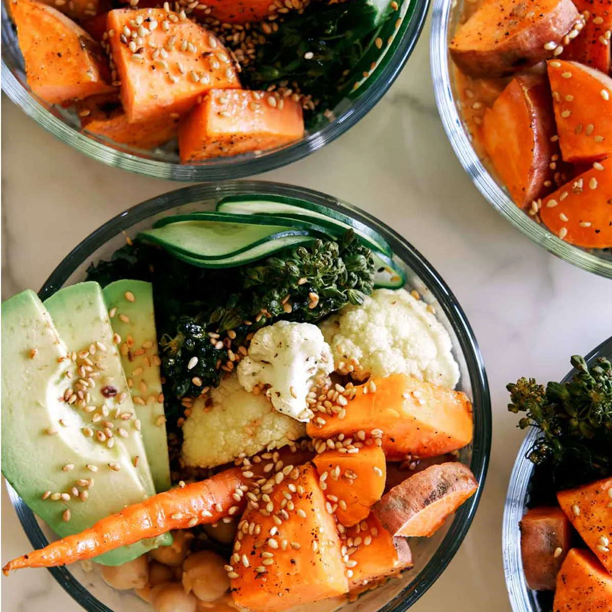 Close-up top-down view of several glass meal-prep bowls arranged on a white marble counter, each filled with roasted sweet potato chunks and whole baby carrots, chickpeas, sliced avocado, cauliflower florets, wilted dark greens and thin cucumber ribbons, all sprinkled with sesame seeds and resting on a base of grains or a creamy spread visible in places — vibrant orange, green and cream tones with varied roasted and fresh textures.