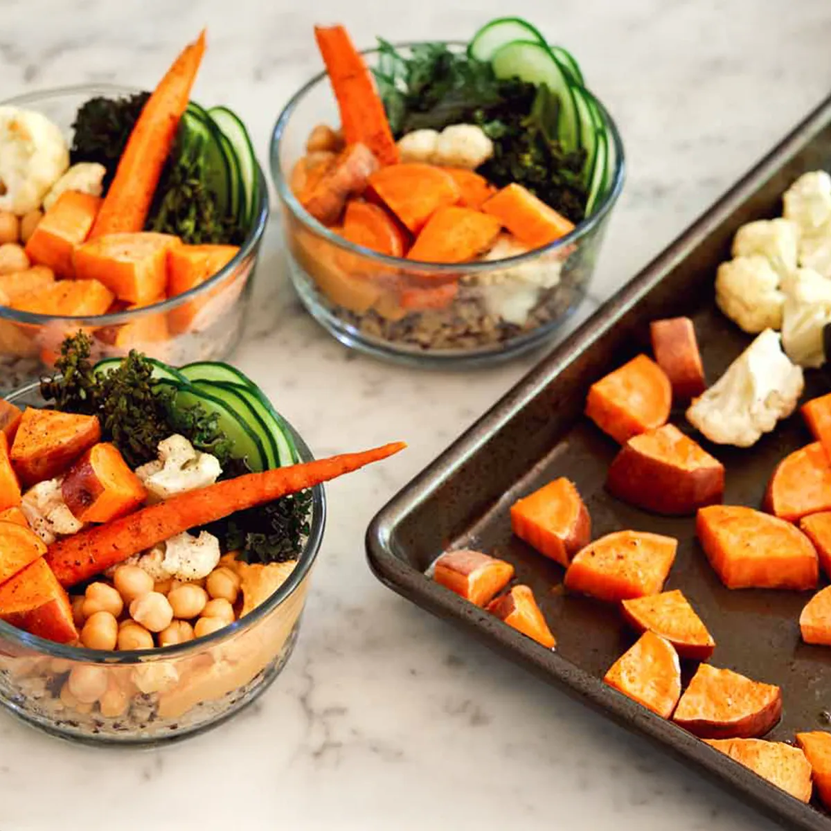 Angled overhead view of several glass meal‑prep bowls on a white marble countertop, each layered with grains and topped with chickpeas, roasted sweet potato chunks, a whole roasted baby carrot, cauliflower florets, wilted dark greens, thin cucumber ribbons and a smear of creamy dressing; a rimmed baking sheet with extra roasted sweet potato and cauliflower sits to the right.