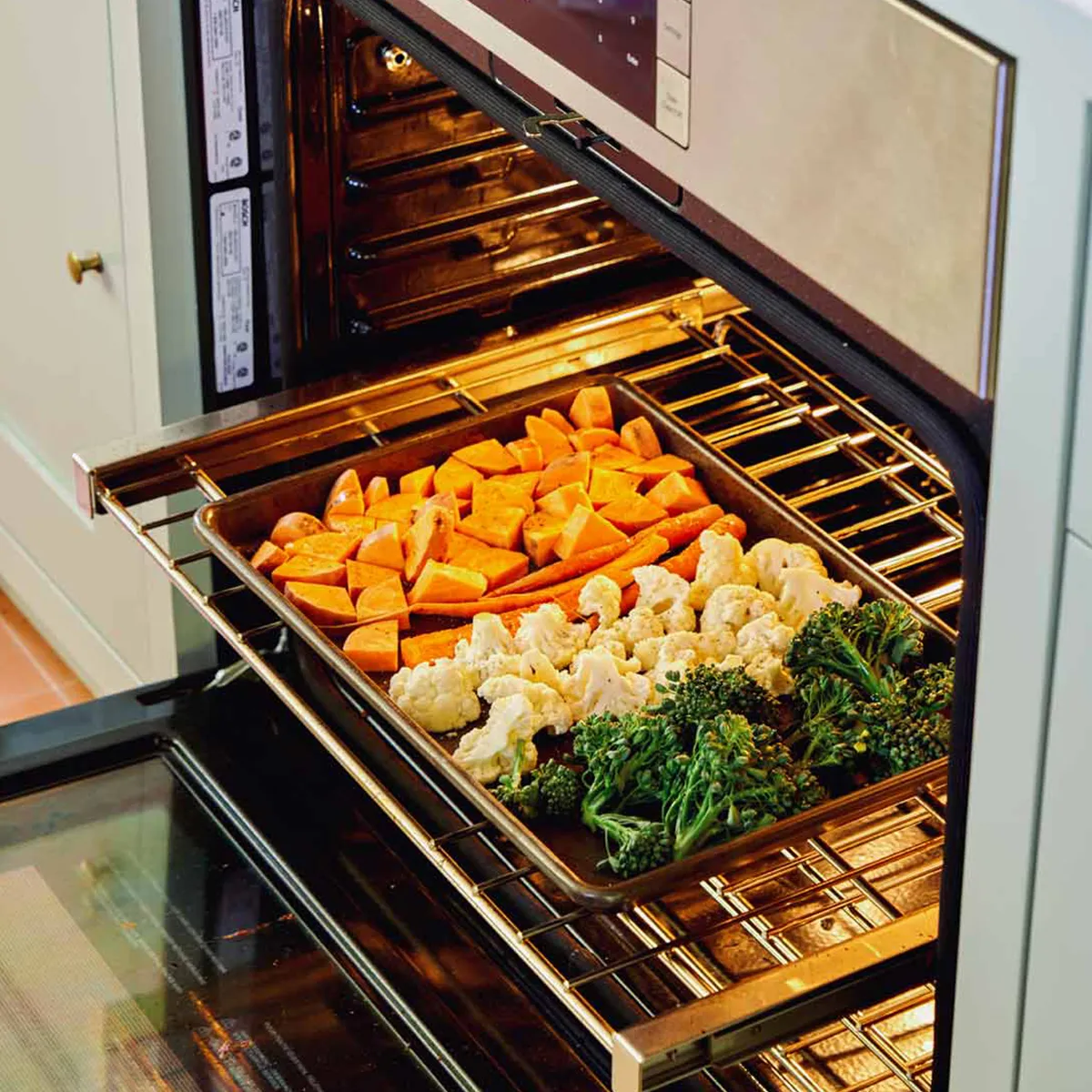 Open stainless-steel oven with a sheet pan of diced sweet potatoes, cauliflower florets, and broccolini roasting on a rack.
