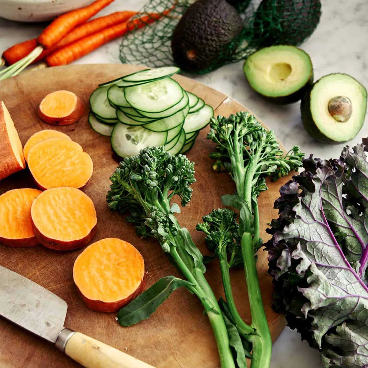 A wooden cutting board with sliced sweet potatoes, cucumbers, broccolini, and kale, surrounded by fresh produce.
