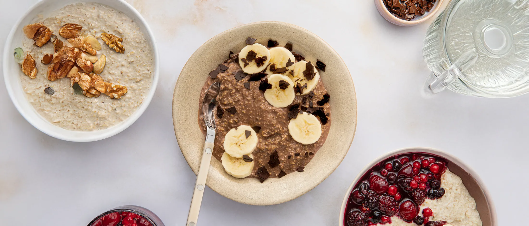 Bowl of chocolate oatmeal topped with banana slices and chocolate shavings, surrounded by bowls of plain oatmeal, mixed berries, and chocolate pieces.
