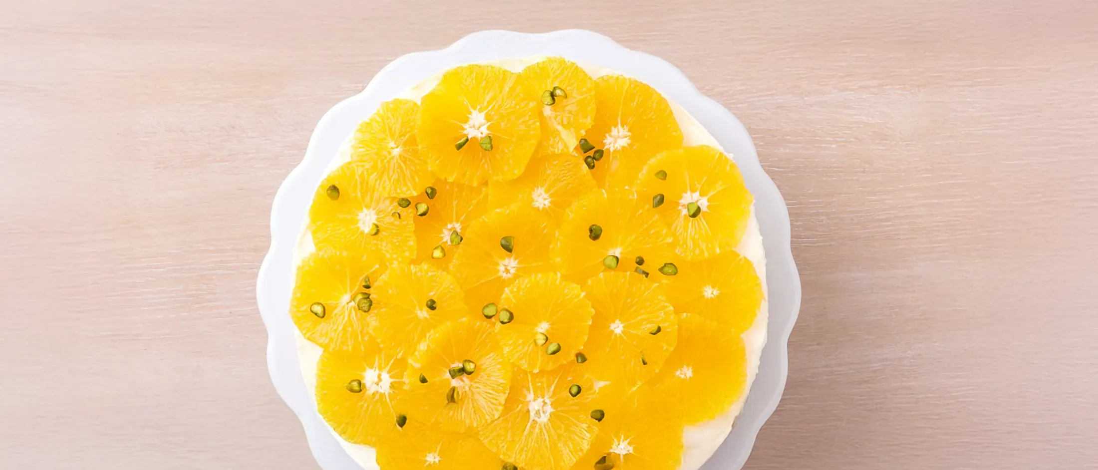 A vibrant orange cake, topped with perfectly arranged orange slices and sprinkled with green pistachios, on a white scalloped plate