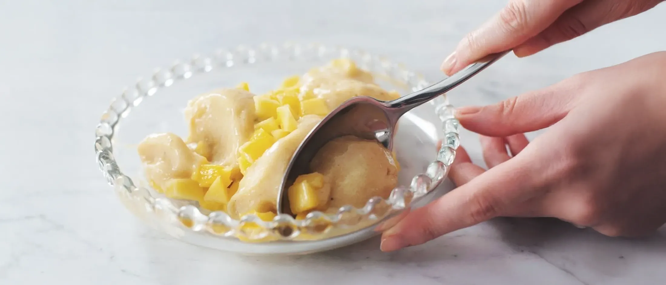Spoon scooping creamy banana mango frozen dessert from a clear glass bowl with beaded edge on a marble surface.