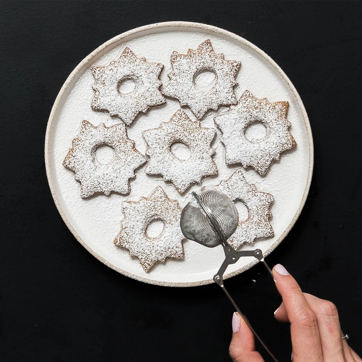 Hand holding a cookie sifter above a plate of star-shaped, powdered sugar-dusted cookies on a black background.
