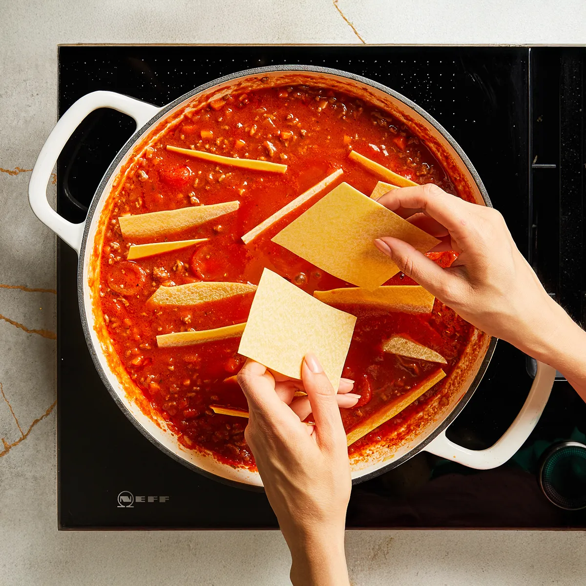 A person places uncooked lasagna noodles into a pot of simmering tomato meat sauce on a stovetop.