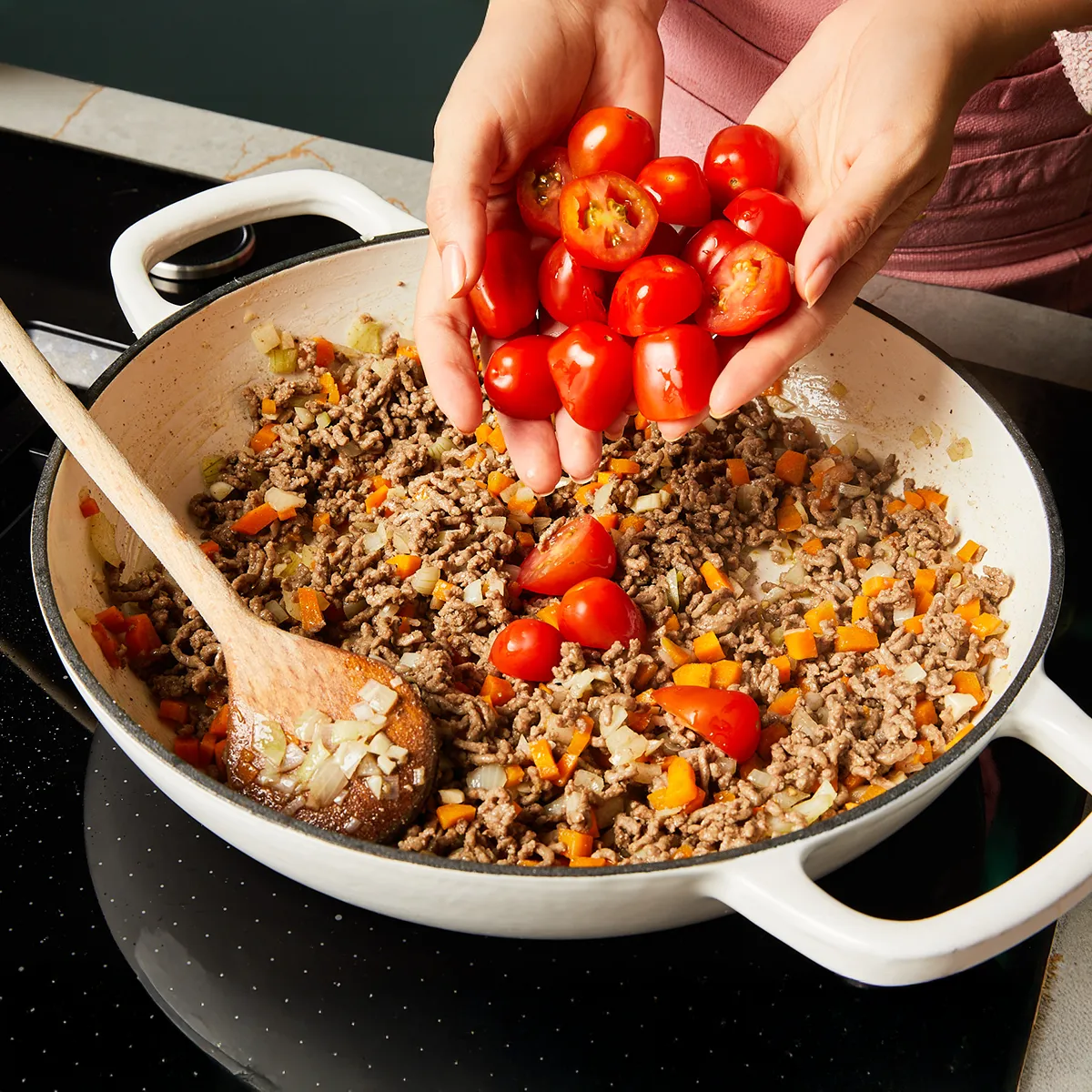 Hands adding halved cherry tomatoes into a pan with cooked ground meat, diced onions, and carrots being stirred with a wooden spoon.