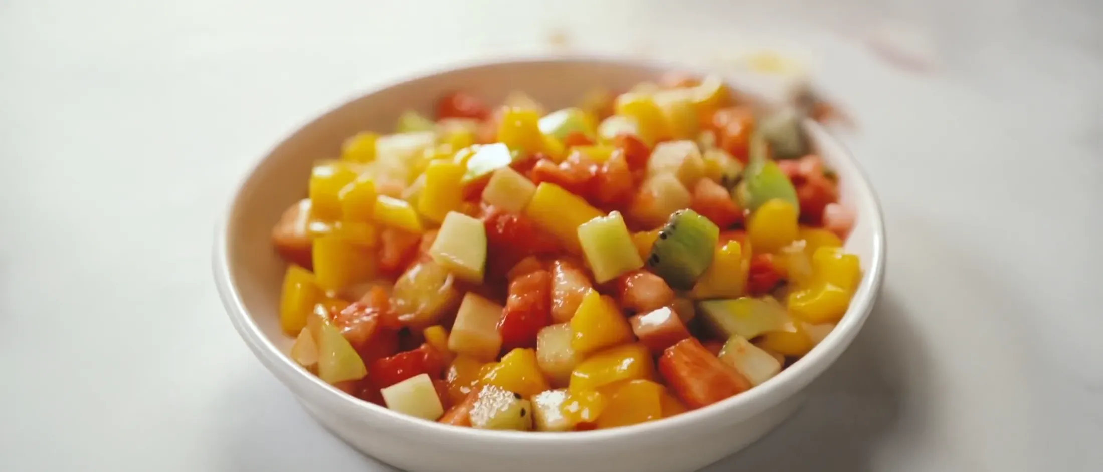 Bowl of mixed fresh fruit salad with diced mango, watermelon, kiwi, and pineapple on a white surface.