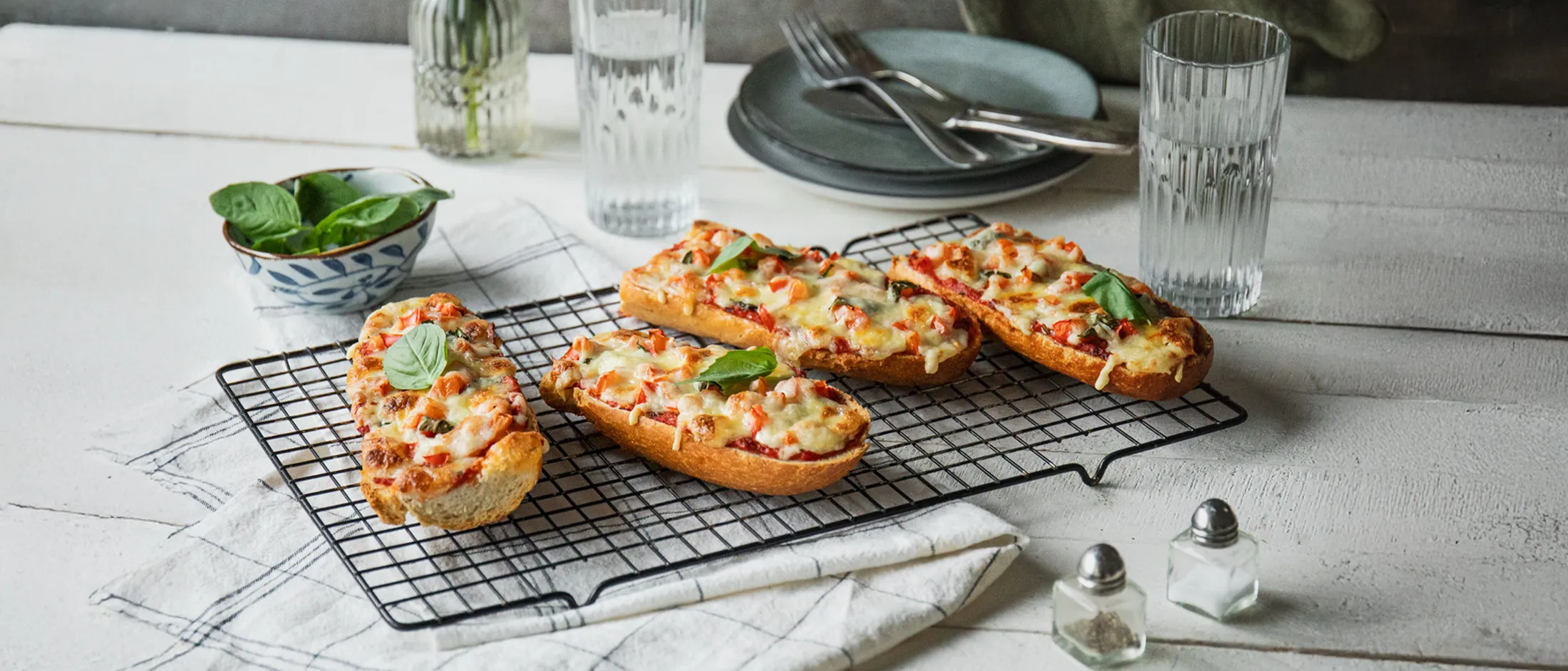 Four slices of French bread pizza with melted cheese and basil leaves on a cooling rack on a white table with glasses, plates, and a bowl of basil.