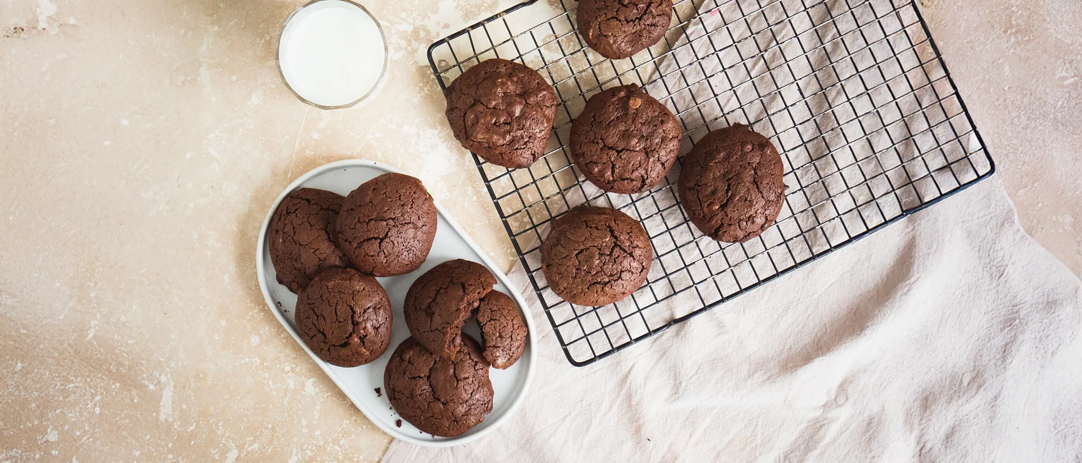 An overhead shot of freshly baked chocolate cookies on a white plate and a cooling rack, with a glass of milk.