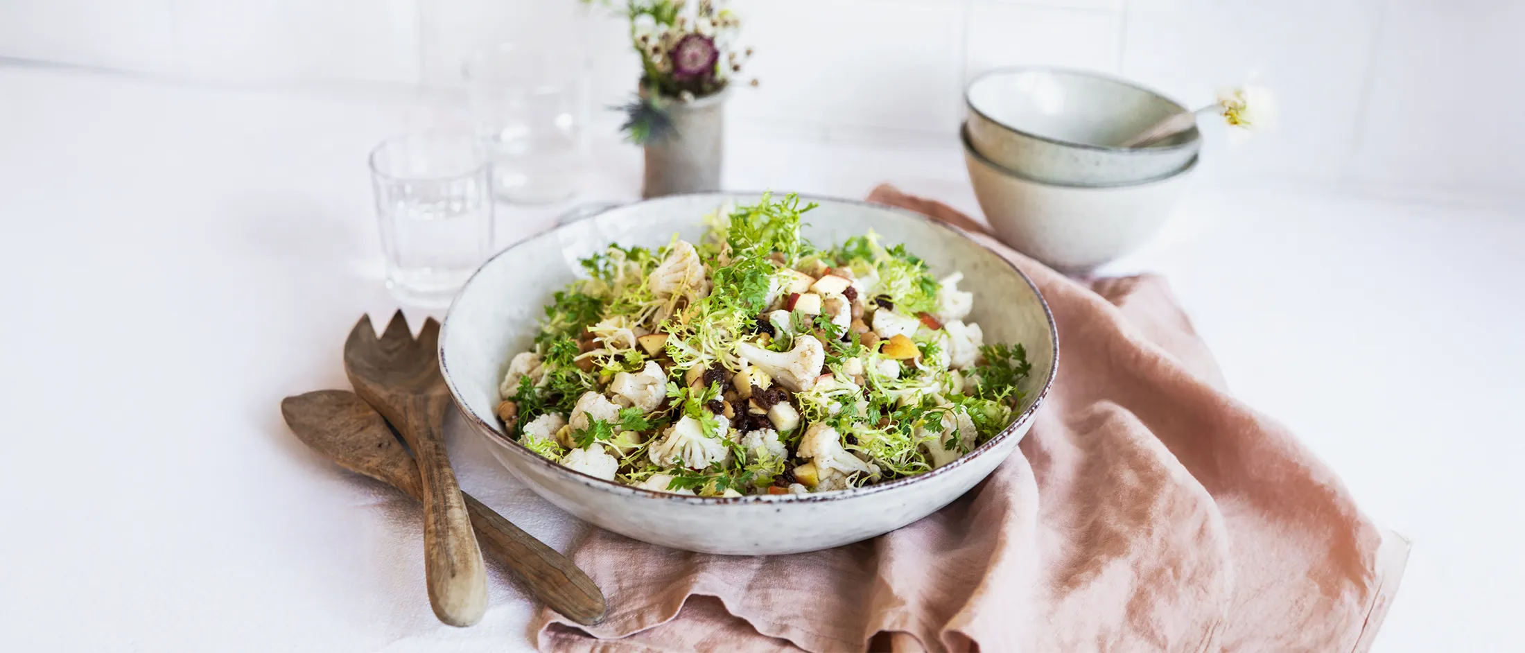 Fresh green salad with cauliflower, herbs, and nuts in a white bowl on a pink cloth with wooden salad utensils nearby.