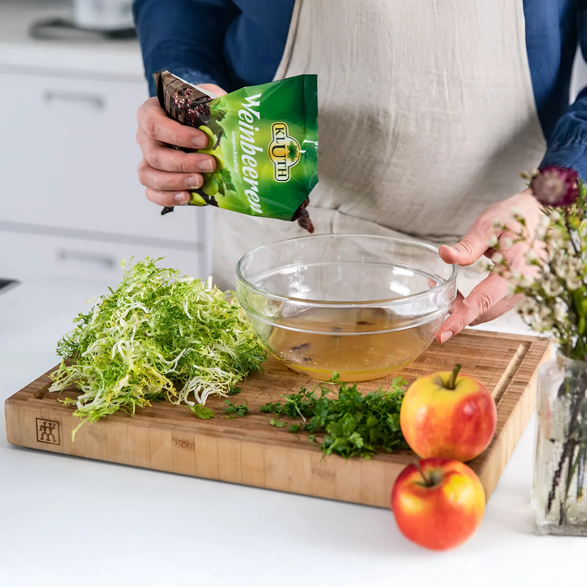 Person pouring soaked dried grapes into a glass bowl on a wooden cutting board with frisée, herbs, and apples nearby.
