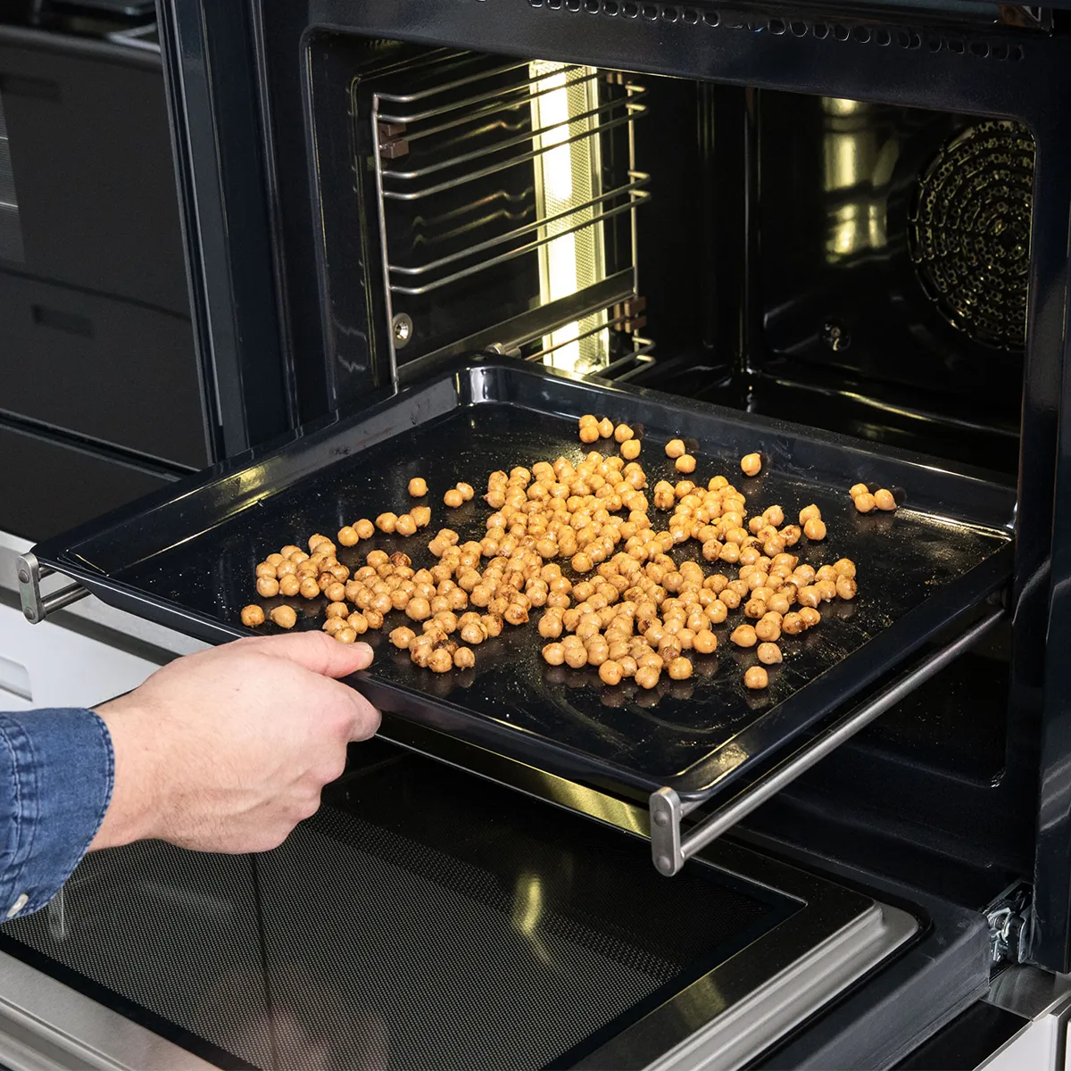 Hand removing a tray of roasted chickpeas from a modern oven in a kitchen setting.