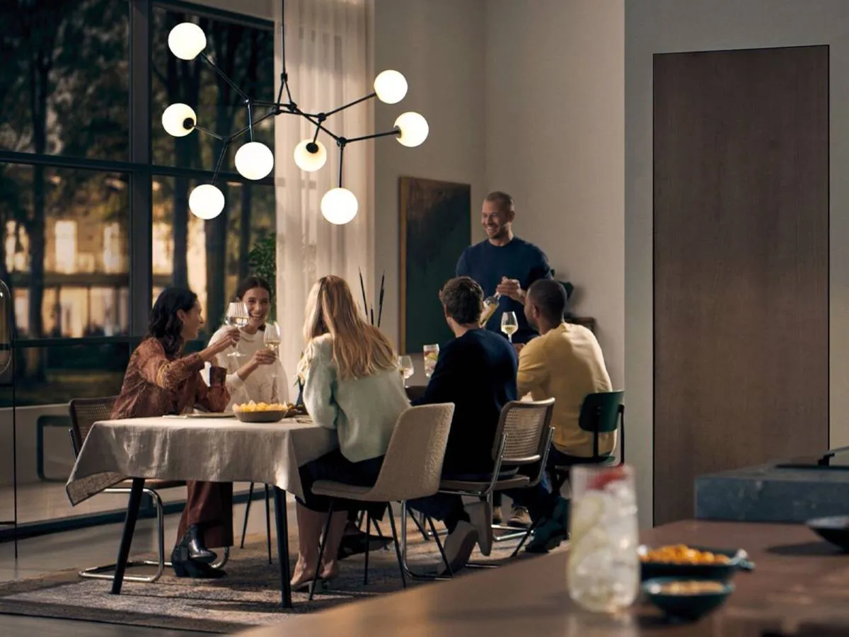 A group of friends enjoys a dinner party in a modern open-plan living space, with a dining table on the left and a sleek kitchen featuring integrated Siemens ovens, a coffee machine, and a microwave on the right.