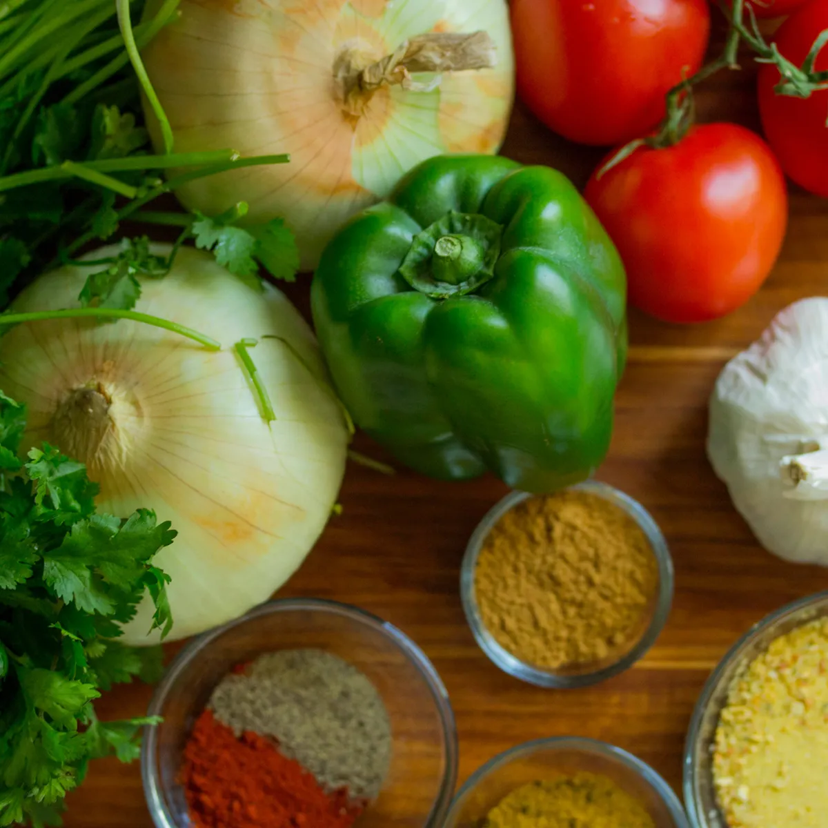 Fresh vegetables including onions, green bell pepper, tomatoes, garlic, cilantro, and bowls of various ground spices on a wooden surface.