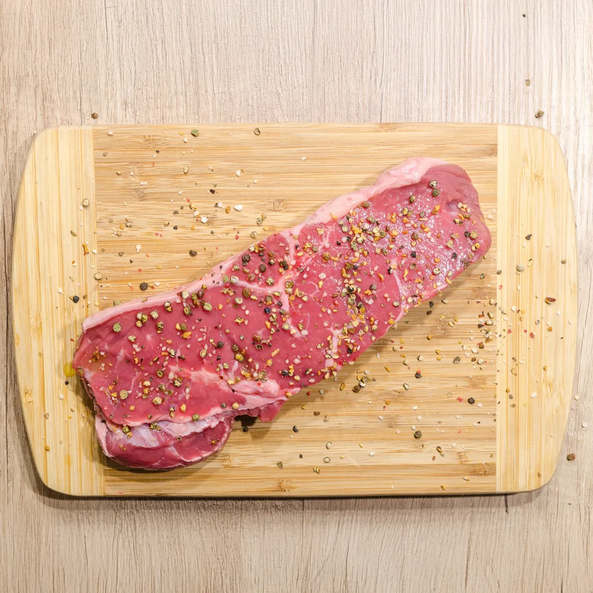 A raw steak heavily seasoned with colorful peppercorns rests on a light wooden cutting board.