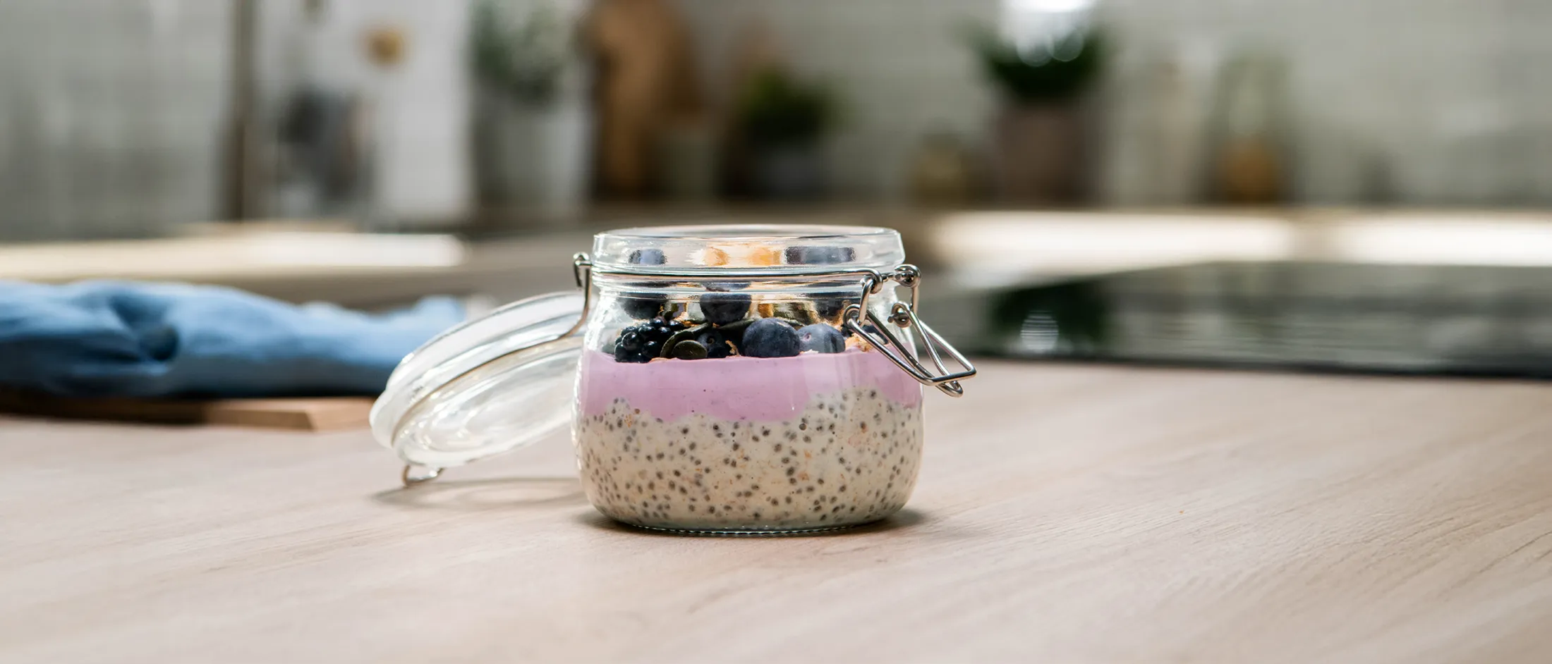 A glass jar containing chia seed pudding layered with pink yogurt and topped with blueberries and granola, set on a kitchen counter.