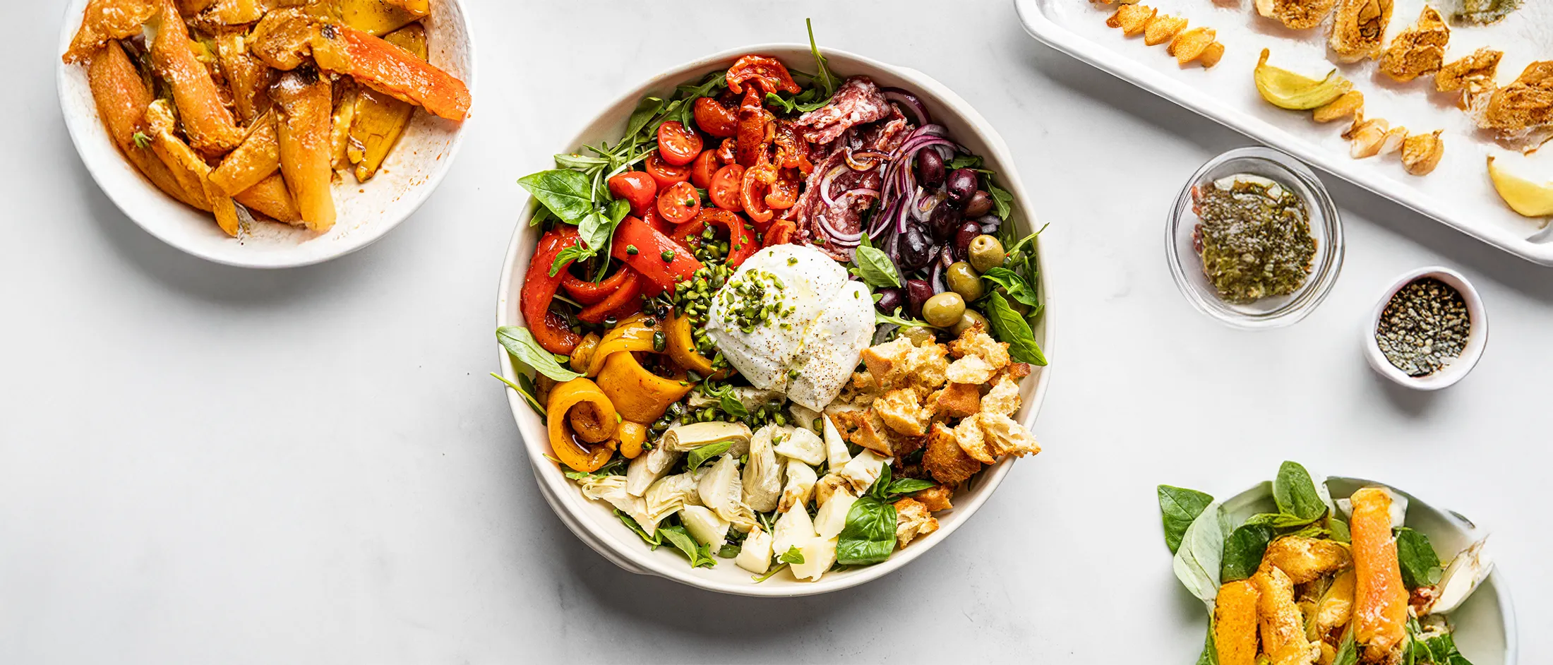 Bowl of Mediterranean salad with mixed greens, roasted peppers, cherry tomatoes, olives, red onion, burrata, artichokes, and toasted bread.