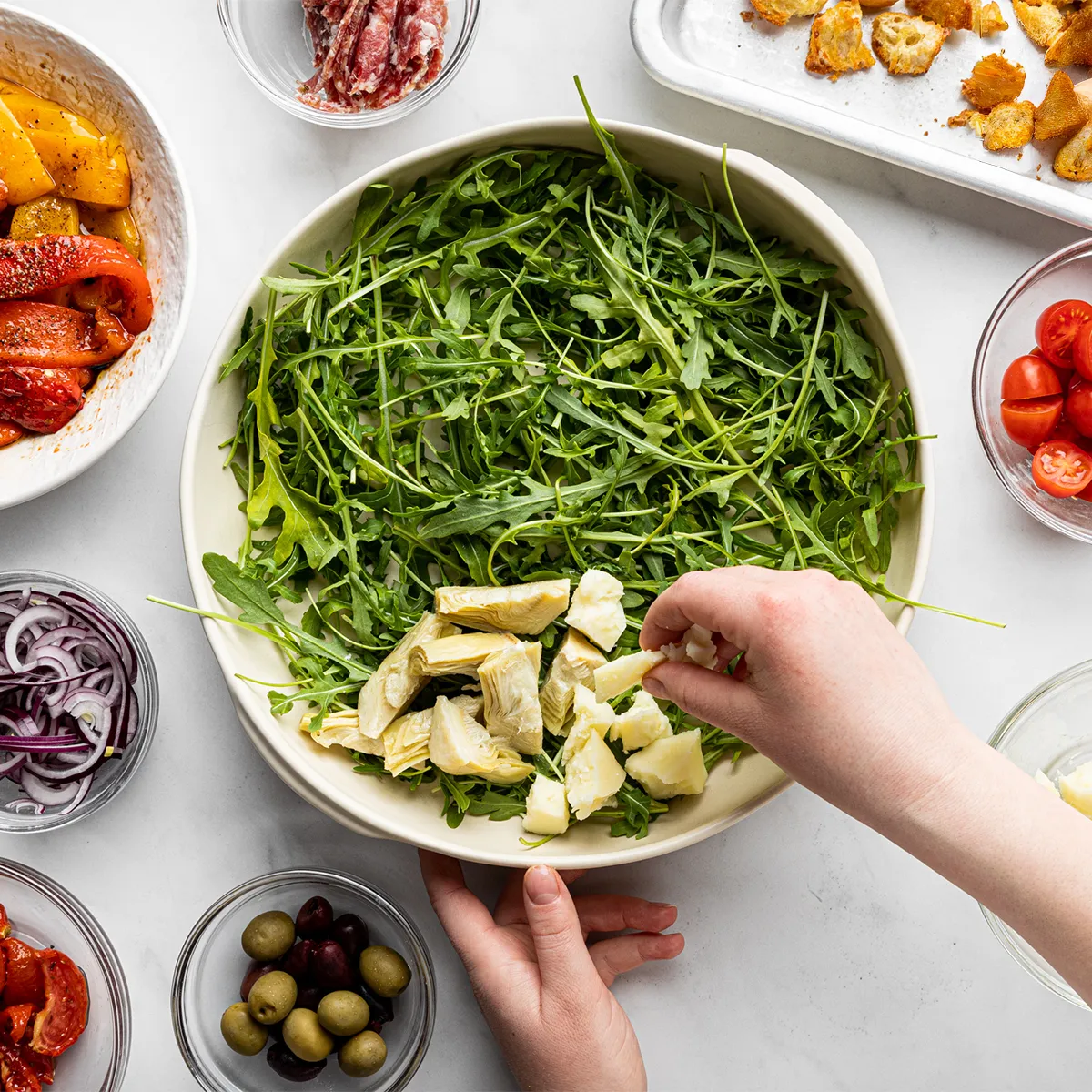 Hands adding mozzarella to a bowl of arugula surrounded by bowls of roasted peppers, olives, cherry tomatoes, and sliced onions.